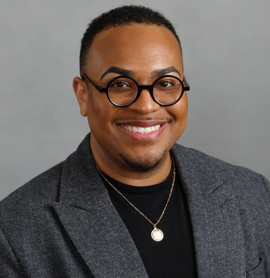 Male in grey suit with black shirt and black round glasses smiling