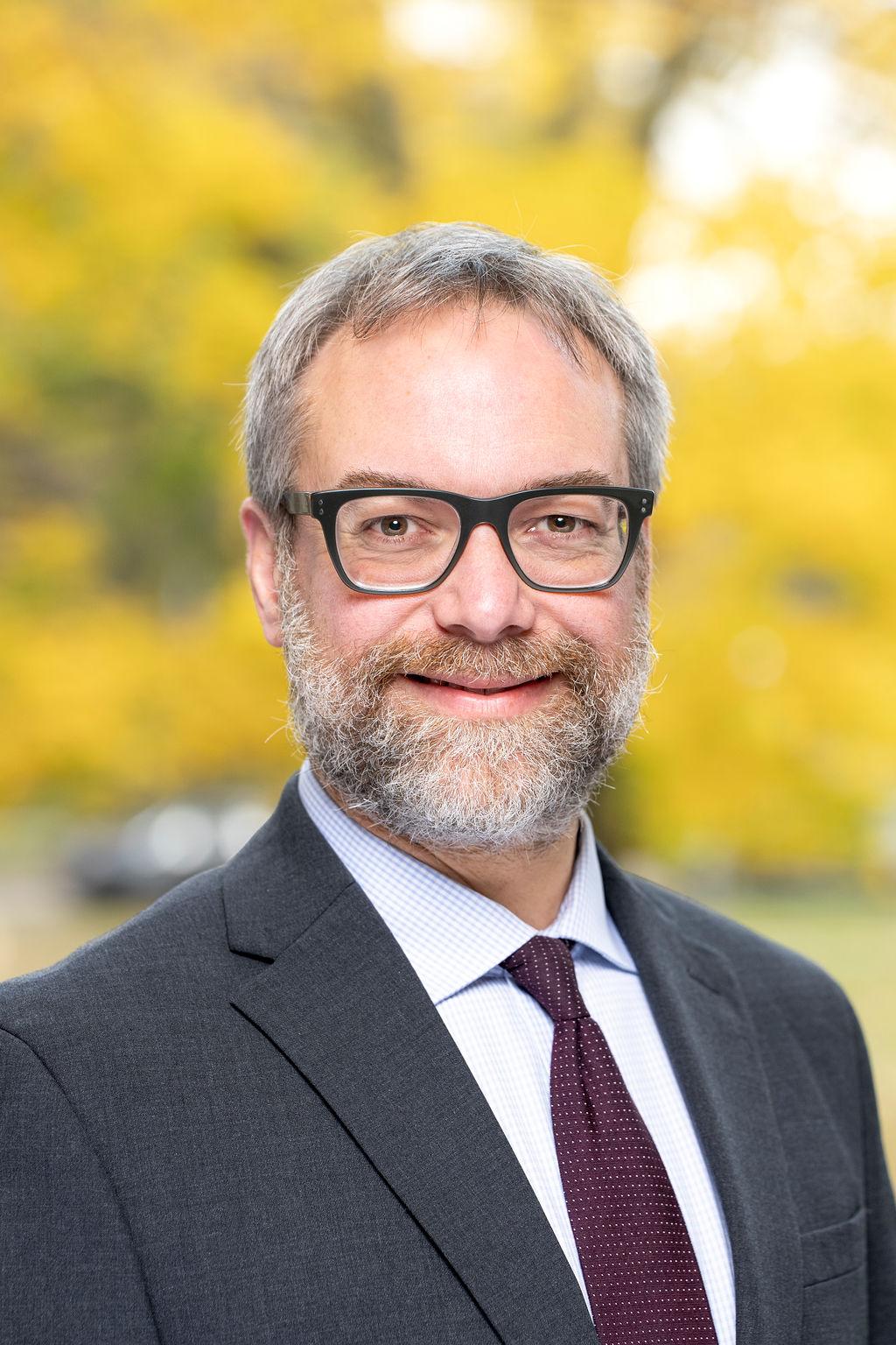 Male in grey suit with maroon tie and white shirt with glasses smiling 