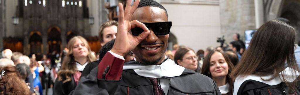 Student in a graduation robe with sunglasses and an hand up symbolizing the okay sign