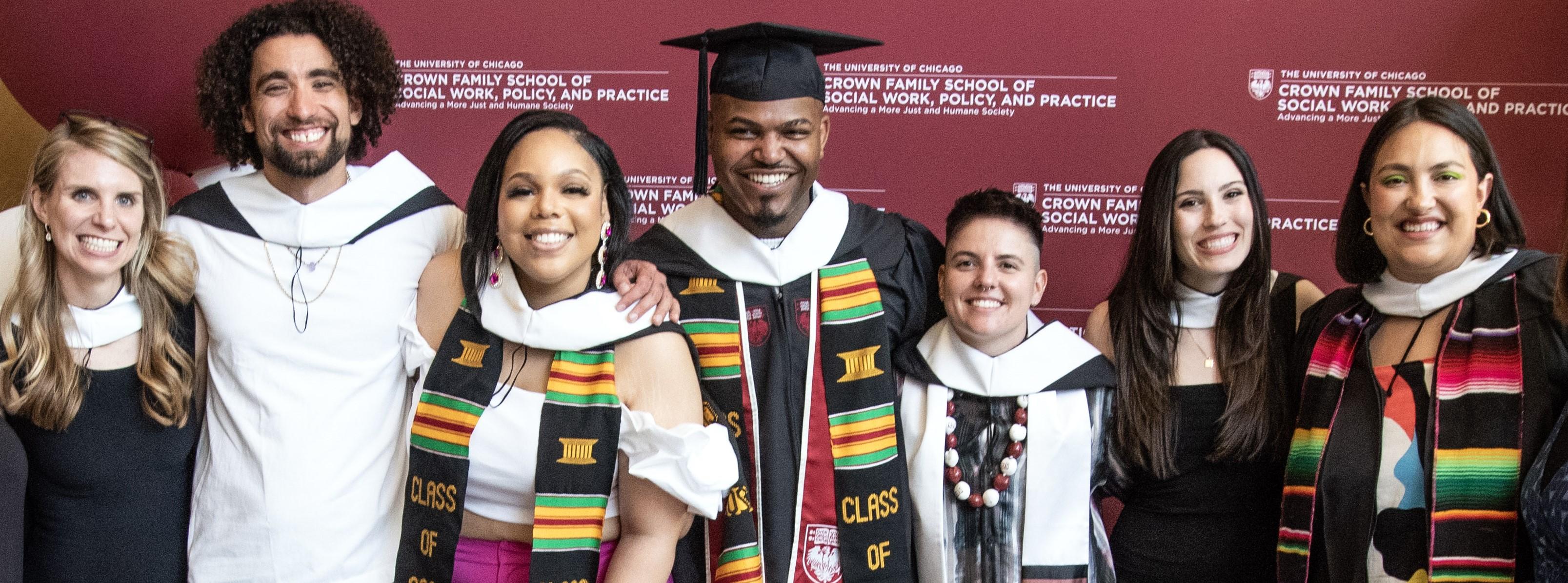 Group of seven students in graduation regalia smiling with arms around each other's shoulders