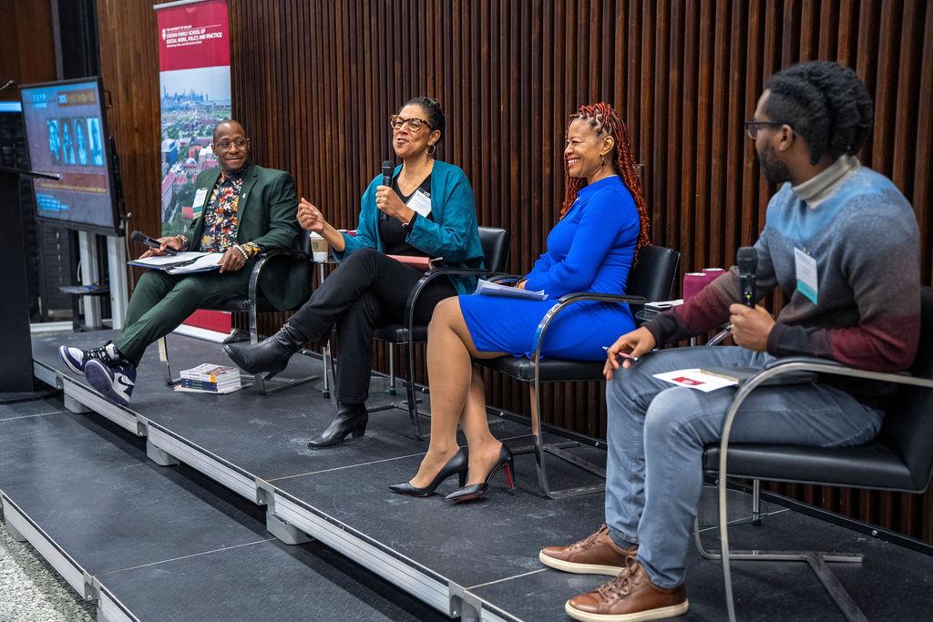 Close shot of the four panelists on stage with woman in blue and black speaking.
