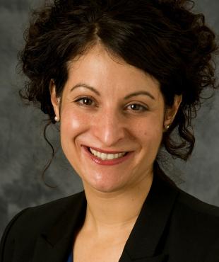 Alida Bouris, a female-presenting person, smiles towards the camera against a dark-gray background.