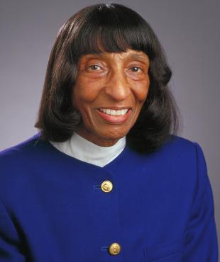 Dolores Norton, a female-presenting person, smiles towards the camera against a light-grey background.