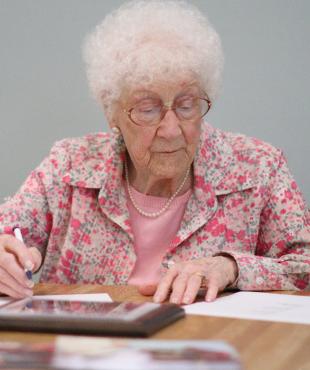 Edythe Kirchmeier seated at a desk signing a paper wearing a pink floral outfit