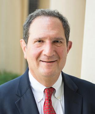 Jerry Wolf wearing a navy suit and red tie against a light background with greenery in the corner