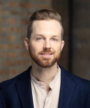 Kevin Barrett wearing a tan shirt under a navy suit jacket against a shadowy tan brick background