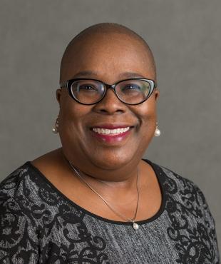 Yolanda Green-Rogers in front of a gray background wearing glasses, pearl earrings, and a dark blouse