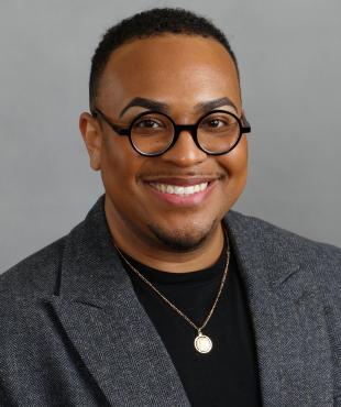 Man in grey blazer with black shirt and black rim glasses