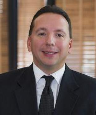 Man in a black suit with white shirt and black tie smiling