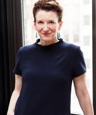 woman standing in a blue dress with hand in pocket by a window with hair pinned up smiling