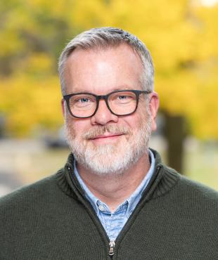 Man in green sweater with blue shirt smiling with glasses and fall leaves background