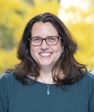 Woman in teal shirt with glasses smiling with yellow trees blurred in background