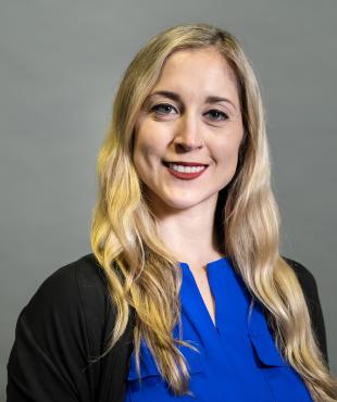 Woman, standing, blue blouse, black sweater, blonde hair , smiling, grey background