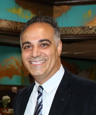 Mane in a black suit with white shirt and black, grey tie smiling in front of colorful background