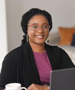 Headshot of female presenting person with black hair loosely pulled black wearing red glasses, a black sweater and fuchsia shirt sitting in front of a laptop.
