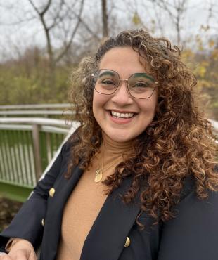 Woman in blue blazer with tan shirt outdoors smiling with glasses and curly hair