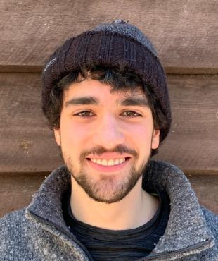Male with grey three quarter zip sweatshirt with black shirt and black and grey winter hat smiling with a brown wood background