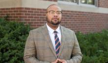Man in brown plaid suit with blue and orange tie standing in front of bushes