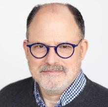 Man with blue glasses blue and white checked shirt with black sweater 