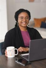 Woman in black blazer with pink shirt sitting at a computer with glasses smiling