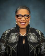 Woman in a black highneck jacket with white star burst and glasses smiling with a short cut and pearl necklace