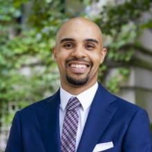 Headshot of Dominque McKoy in a blue suit with pink and blue tie outdoors smiling