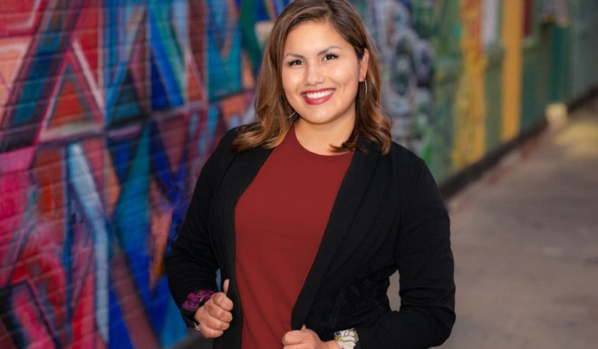 woman in a black jacket with maroon top standing in front of a mural wall smiling