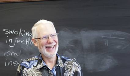 A male-presenting person laughs while facing away from the camera in front of a chalkboard.